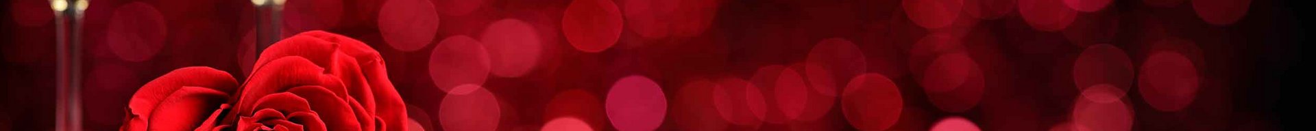 Et hjerteformet rødt rose ligger på en hvid tallerken med bestik ved siden af. I baggrunden ses to glassene fyldt med gylden drink, alt på en rød baggrund. Billedet giver en romantisk stemning.